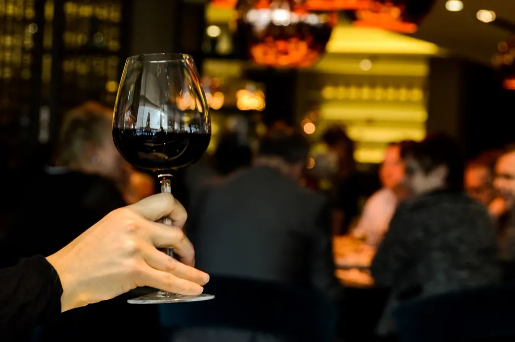 A glass of red wine raised in a busy, warmly lit bar